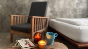 Una cama, una silla, una mesa con una vela, una taza y un cuadro enmarcado.