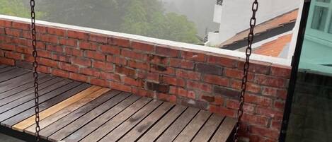 Un banco de madera con un fondo de pared de ladrillo, una cadena colgante y vista de un edificio a través de una ventana.