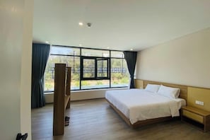 A bedroom with a large bed, a wooden floor, a window with curtains, and a nightstand.