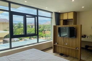 A bedroom with a large window, a bed, a TV mounted on a wooden cabinet, and a small desk area.