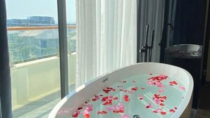 A modern bathroom with a freestanding bathtub filled with rose petals, large windows, and a view of a cityscape. - Aurora Bao Anh Hotel (Binh Kien)