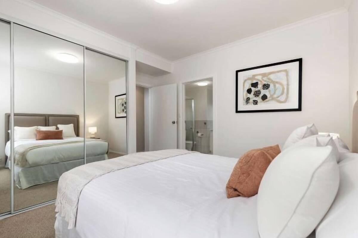 Habitación de hotel con una cama grande, un espejo, una obra de arte abstracta enmarcada y un baño separado visible a través de una puerta de cristal.