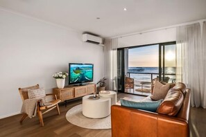 A modern living room with a television, seating, a round coffee table, and a large window with a view of the sea. - Sol Sands: Oceanfront Balcony Henley Beach (Henley Beach)