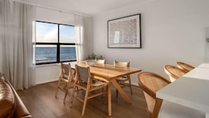 A modern living room with a wooden dining table, chairs, a sofa, and a large window offering a view of the sea. - Sol Sands: Oceanfront Balcony Henley Beach (Henley Beach)