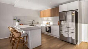 A modern kitchen with white cabinetry, a stainless steel refrigerator, a built-in oven, and a wooden island with bar stools. - Sol Sands: Oceanfront Balcony Henley Beach (Henley Beach)