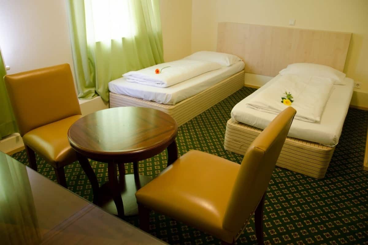Hotellihuone, jossa on kaksi erillistä sänkyä, pyöreä puinen pöytä, kaksi tuolia ja vihreäkuvioinen matto.