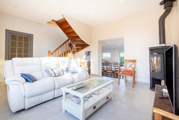 A living room with a white sofa, a glass coffee table, a wooden staircase, and a wood-burning stove.
