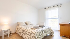 Ein Schlafzimmer mit Bett, Nachttisch, Lampe und Blick ins Freie durch ein Fenster.