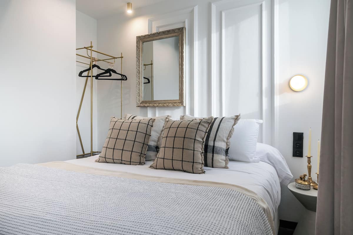 A neatly arranged bedroom with a bed, pillows, a mirror, a bedside table with candles, and a clothing rack.