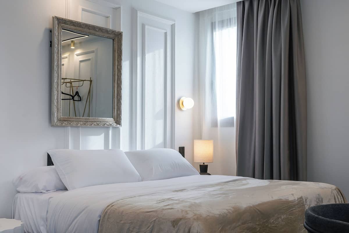A hotel room with a large bed, a mirror, a bedside lamp, and a view of the outdoors through a window with curtains.