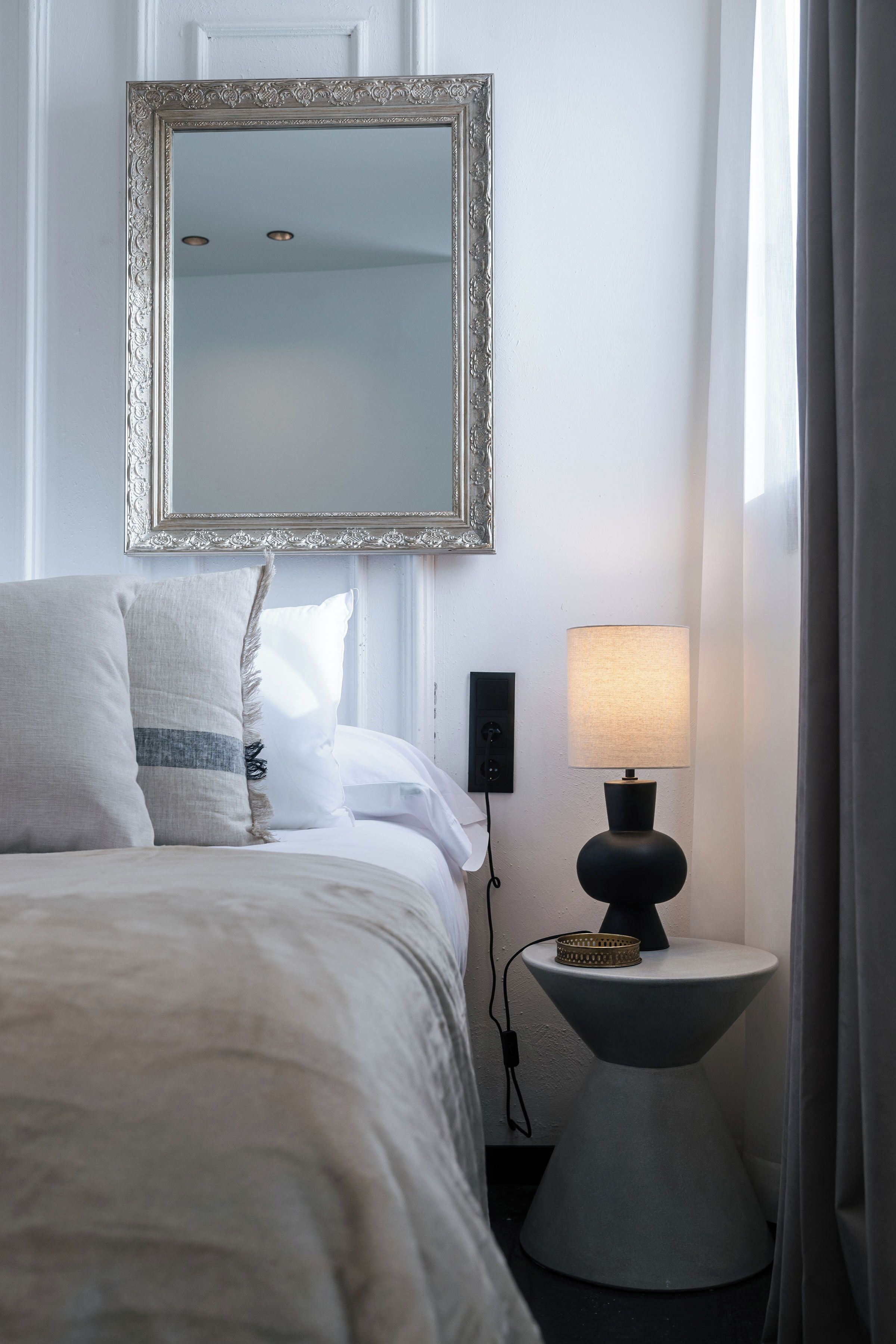 A neatly made bed with a white comforter and pillows, a bedside table with a lamp, and a large mirror on the wall.