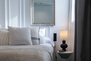 Ein ordentlich bezogenes Bett mit weißer Bettwäsche und Kissen, ein an der Wand befestigter Spiegel, ein Nachttisch mit Lampe und eine Steckdose.