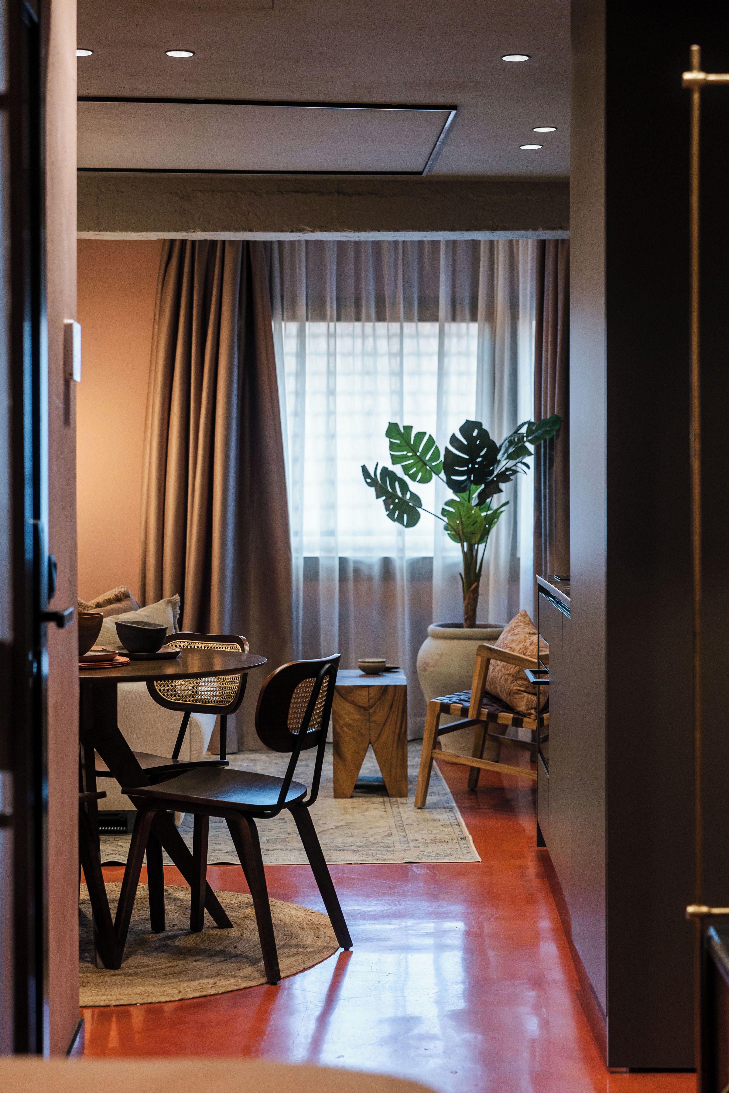 Un espacio habitable compacto con zona de comedor, una planta grande en maceta y vistas a través de una ventana con cortinas.