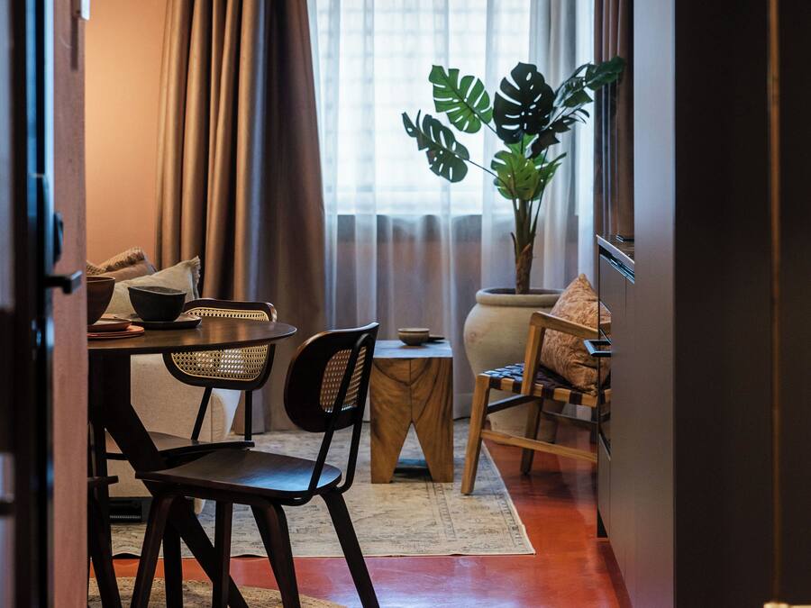 Un espace de vie compact comprenant un coin repas, une grande plante en pot et une vue sur l’extérieur à travers une fenêtre avec des rideaux.