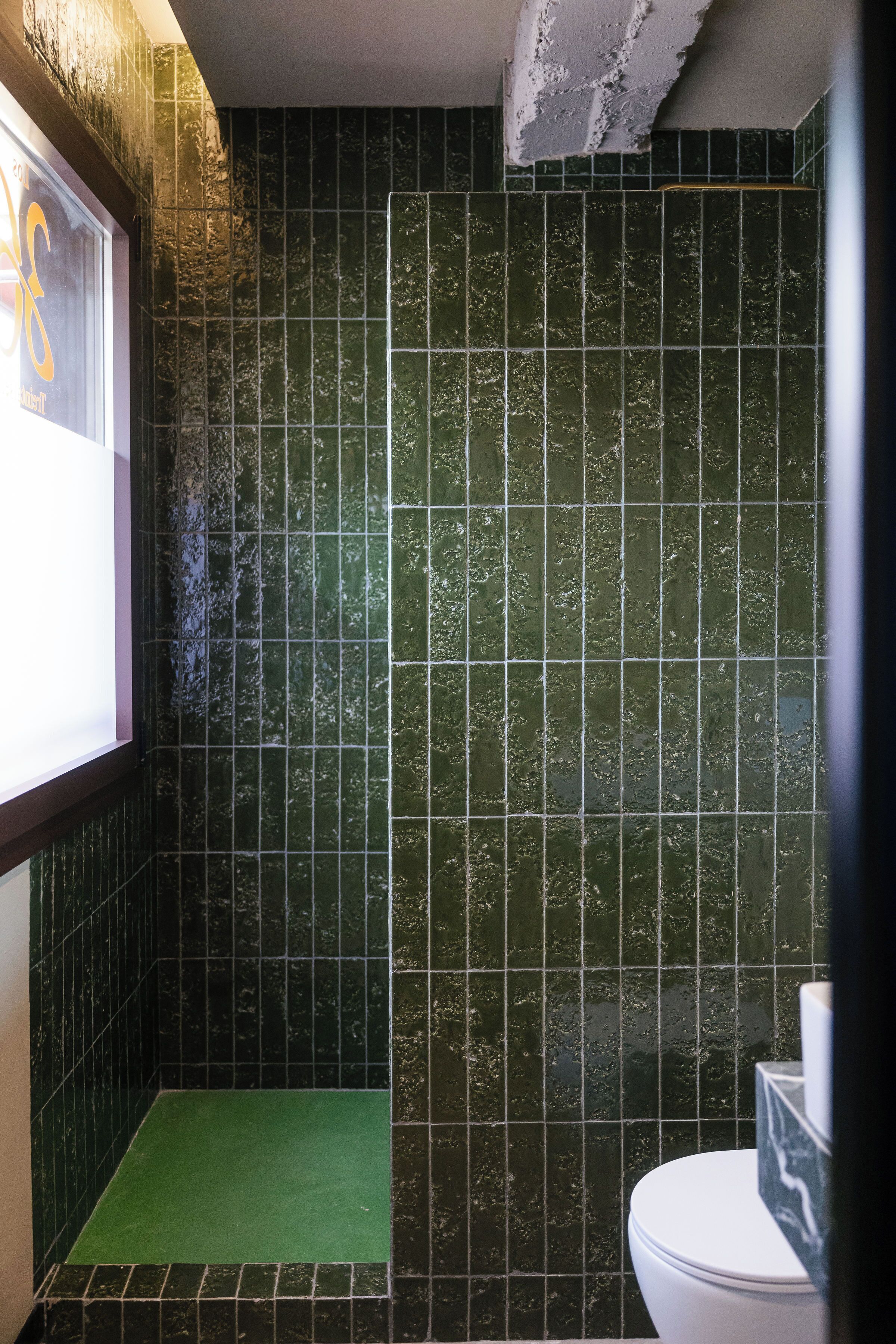 A bathroom with dark green tiles, a white toilet, and a green mat.