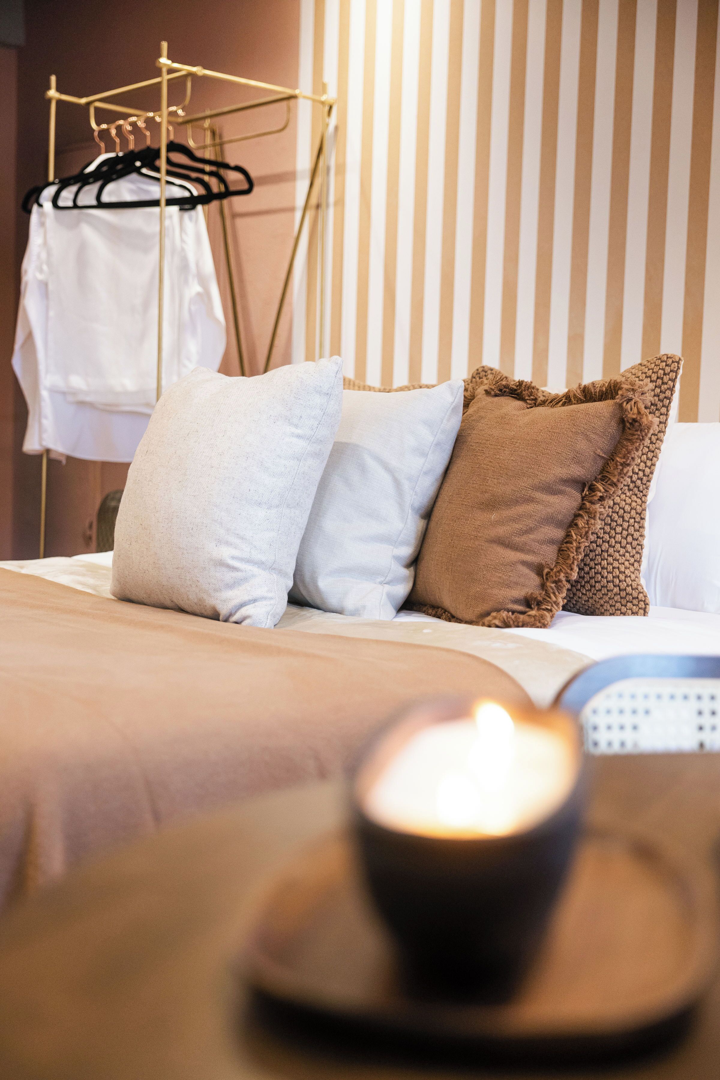 A neatly made bed with pillows, a hanging clothes rack, and a lit candle on a tray.