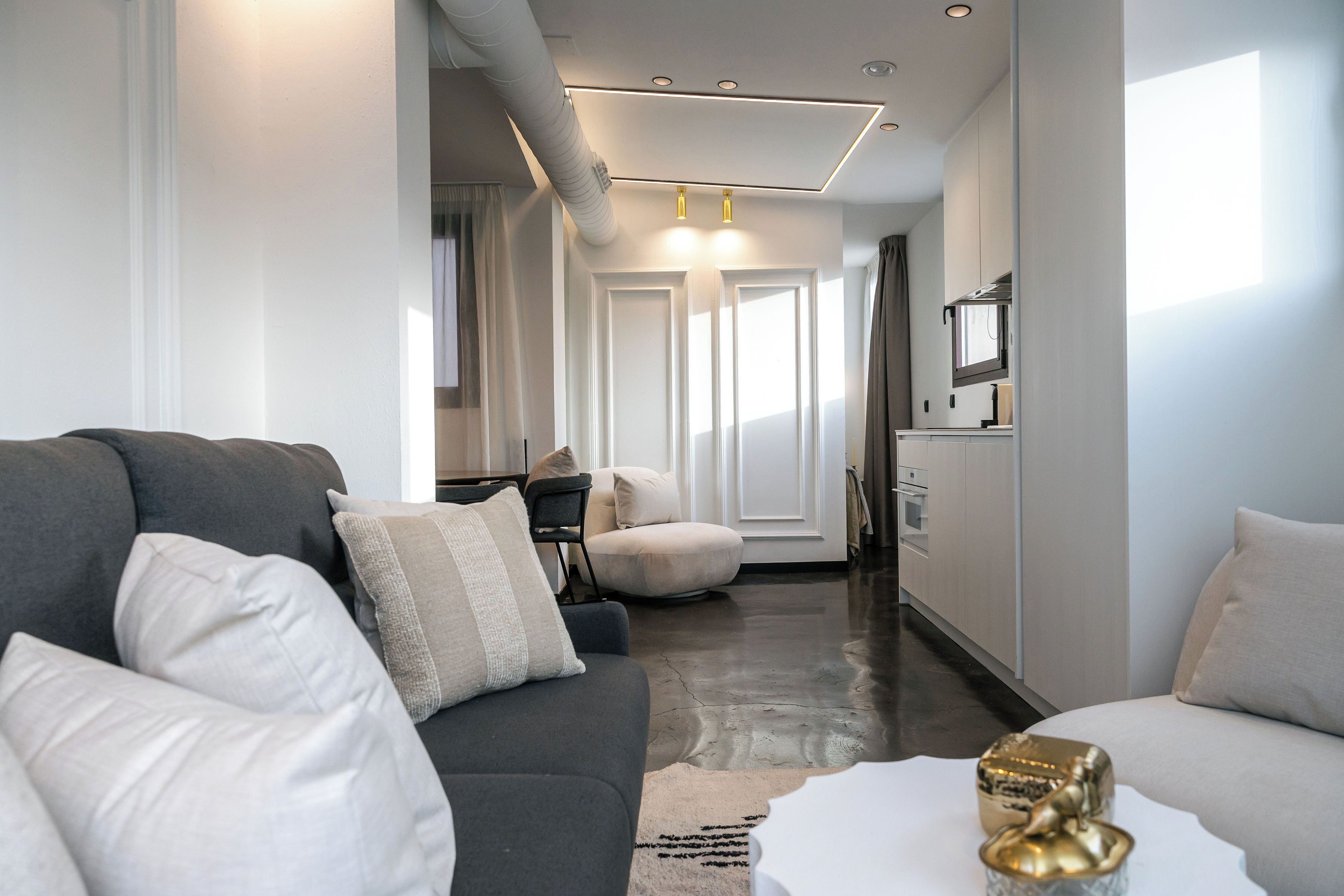 A modern living room with a grey sofa, a round ottoman, and a white coffee table. There is a kitchen area visible in the background with a window and a cabinet.