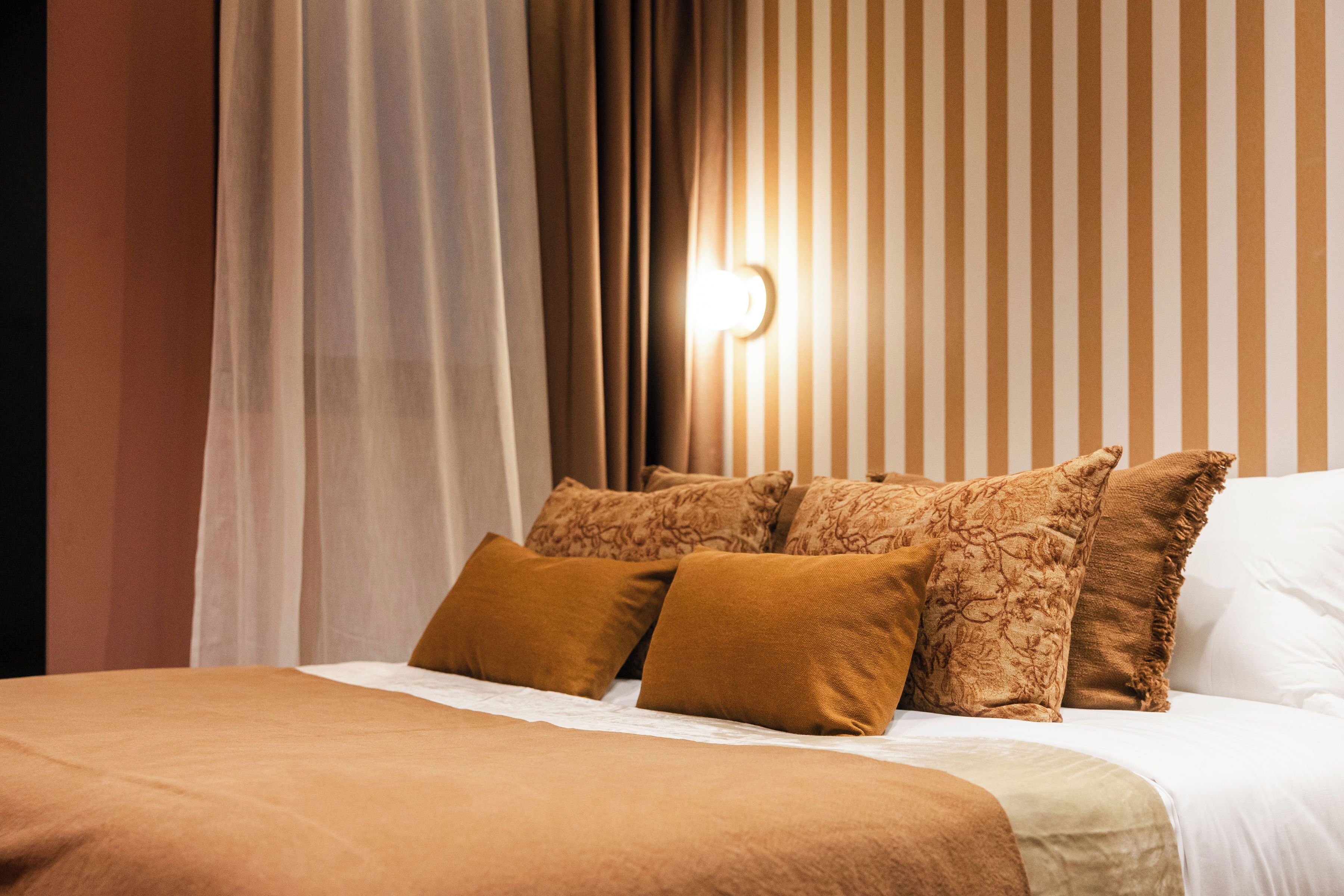 Una cama bien hecha con ropa de cama y almohadas marrones, un pared a rayas al fondo y una fuente de luz en la pared.