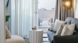A modern living room with a grey sofa, white armchair, and a small round coffee table. A large window with sheer curtains allows natural light to fill the space. - LOS 36 BY GRUPO CCOMMO (Murcia)