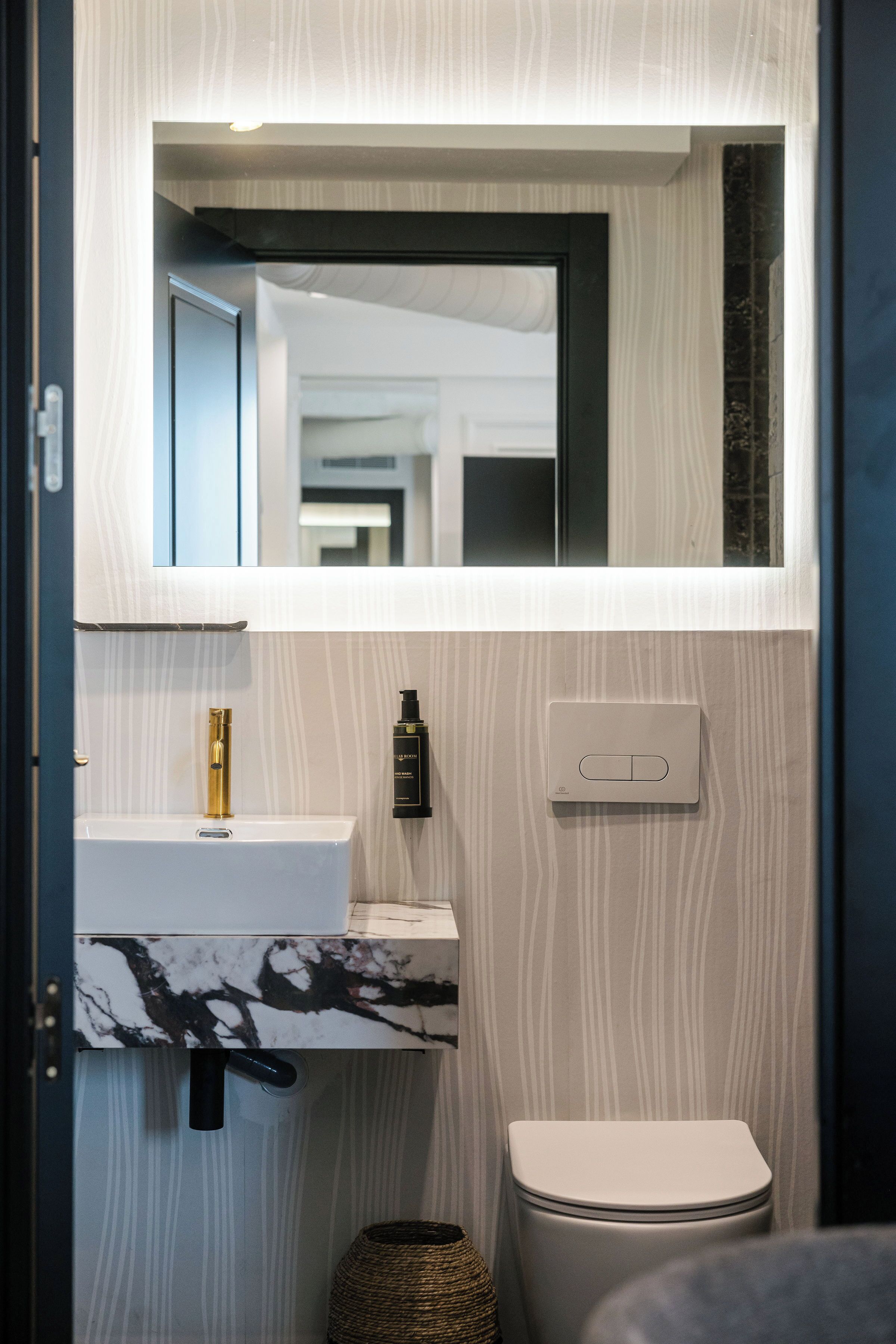 Ein Badezimmer mit Toilette in Weiß, Marmorwaschbecken, Goldarmatur, einer schwarzen Flasche und einem Lichtschalter.