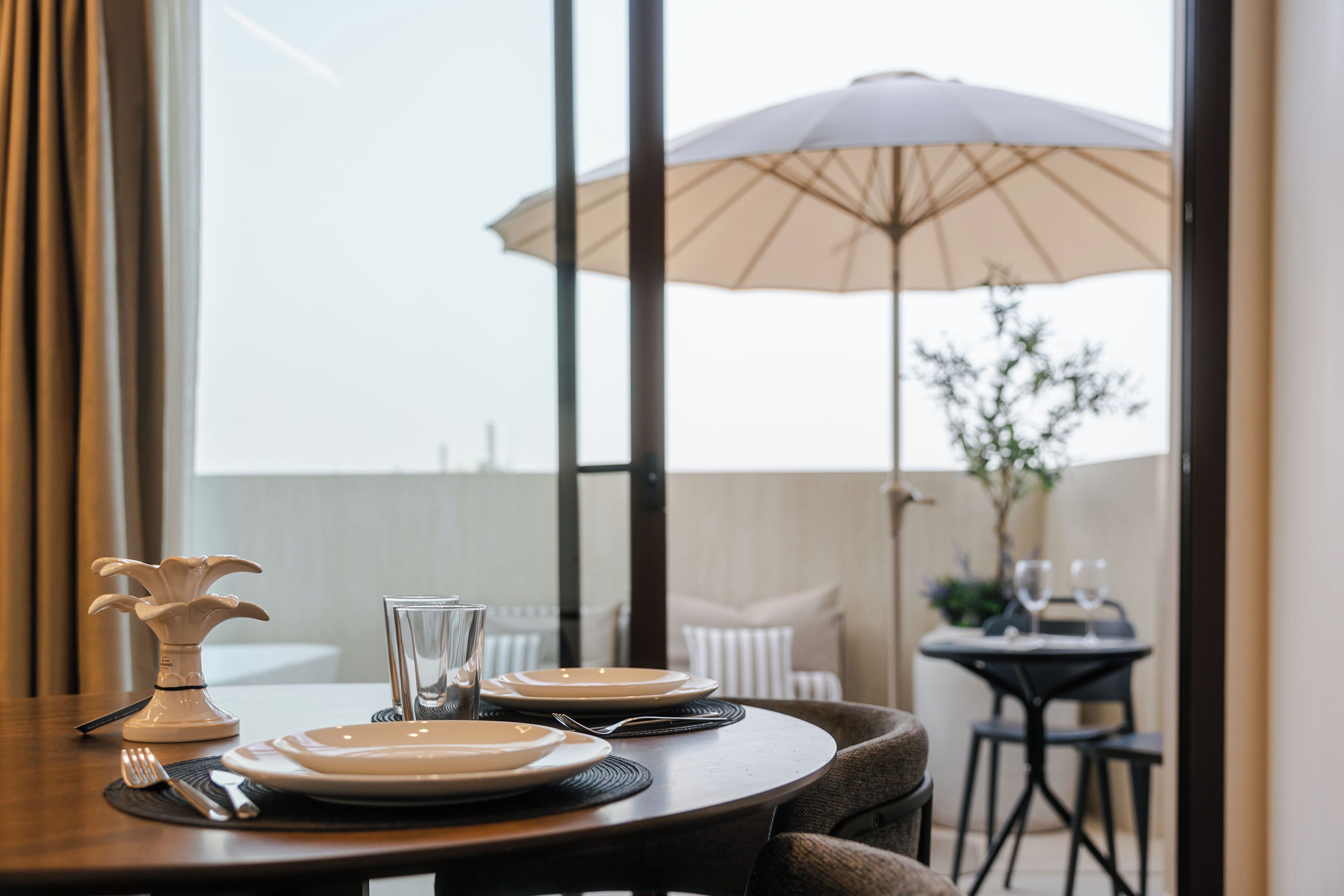 Una mesa de comedor dispuesta con platos, cubiertos y una copa, con una sombrilla y una planta a la vista al fondo.