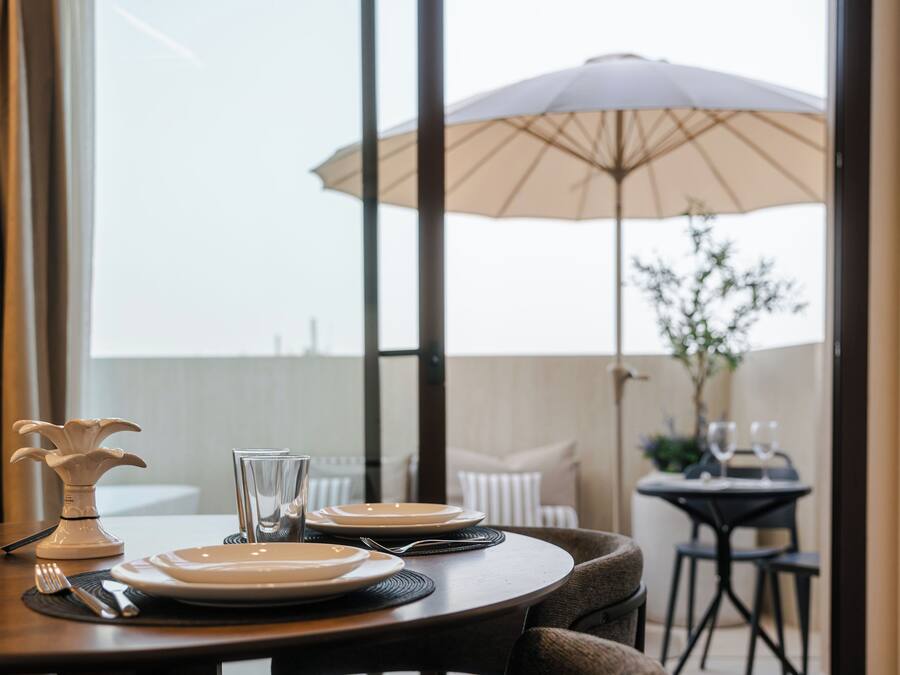 Una mesa de comedor dispuesta con platos, cubiertos y una copa, con una sombrilla y una planta a la vista al fondo.