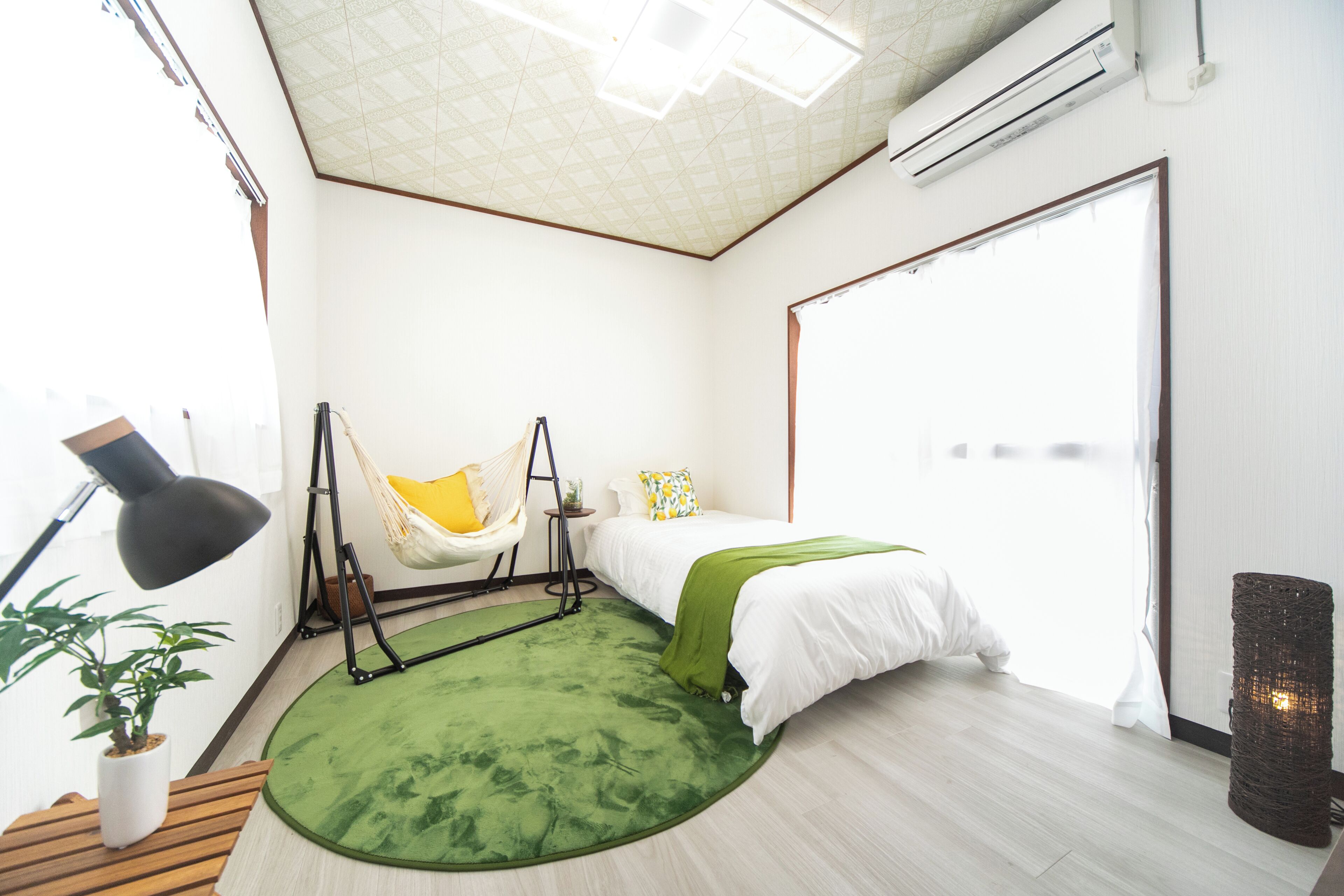 A modern bedroom with a bed, a green rug, a hanging chair, a potted plant, and an air conditioning unit.