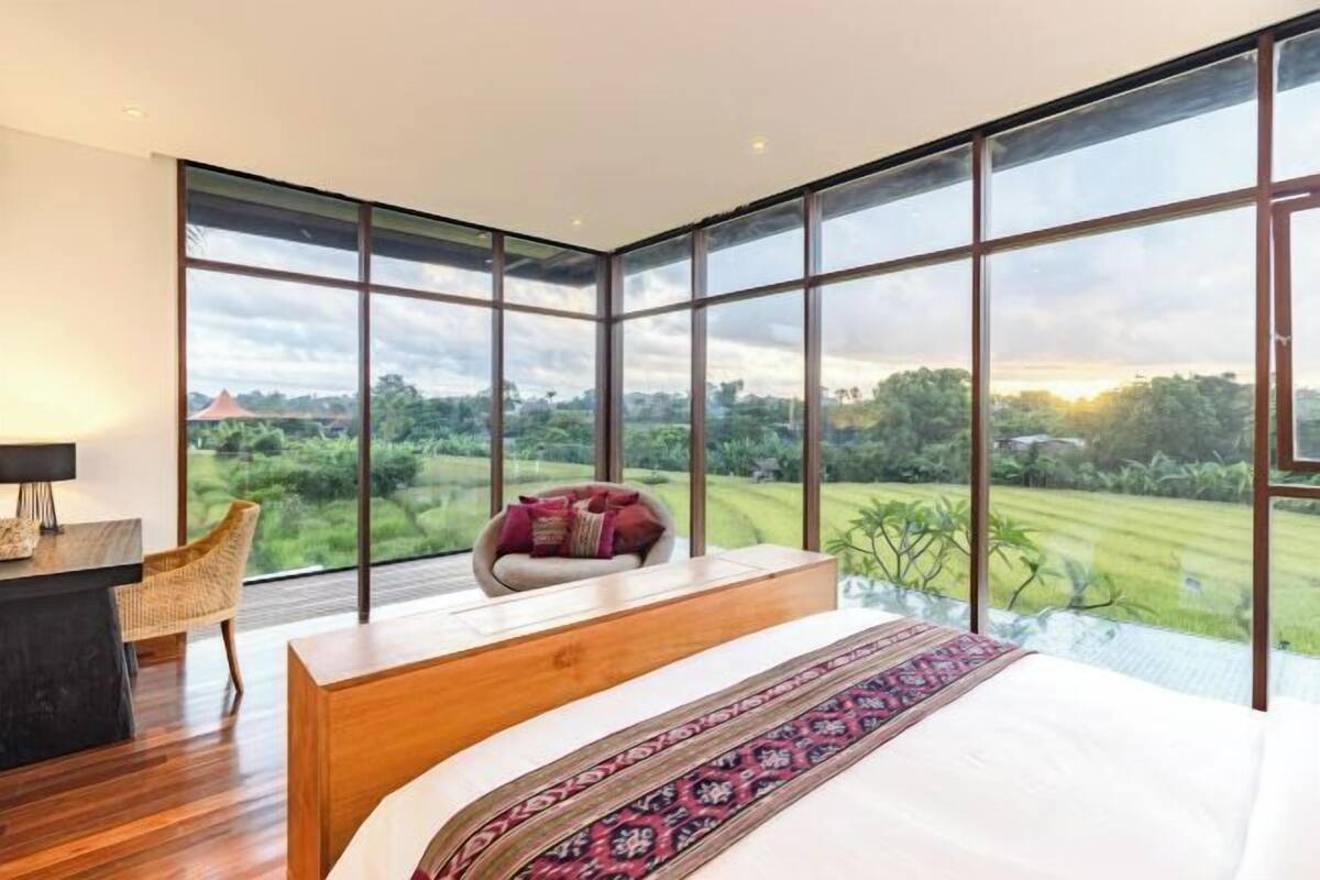 A modern bedroom with a large bed, a desk, and a chair, featuring a large window with a view of a green landscape.