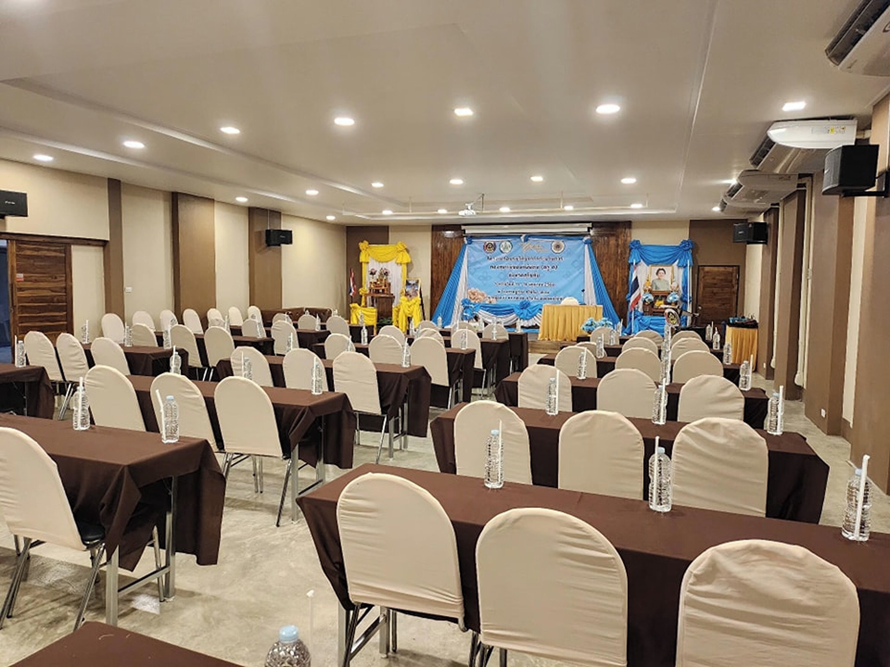 Un salón de conferencias con filas de mesas y sillas, un escenario con un cartel y un atril.