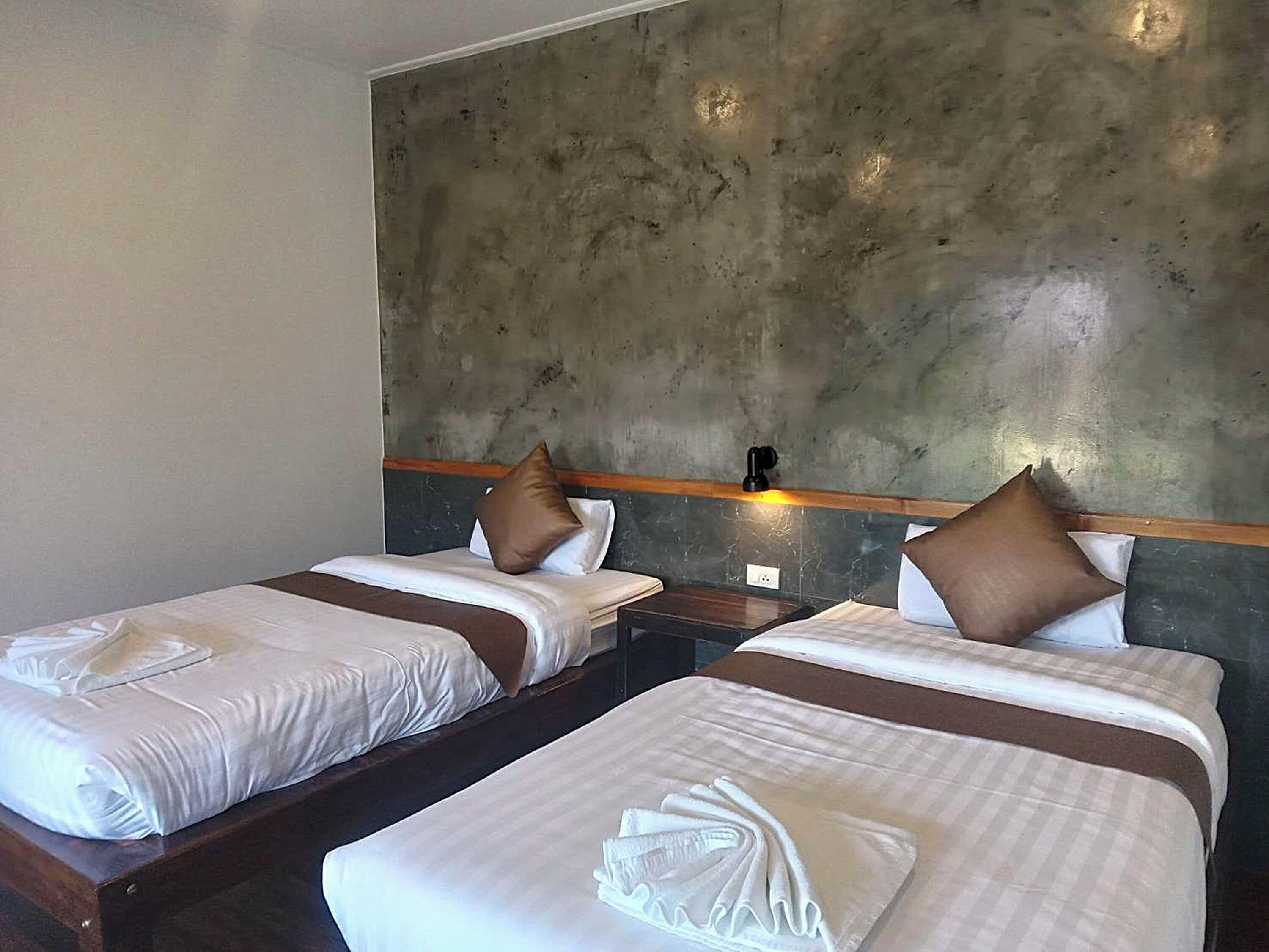 Une chambre d’hôtel avec deux lits simples, chacun doté de draps blancs et d’oreillers marron. La chambre est agrémentée d’un mur d’accent en béton et d’une tête de lit en bois.