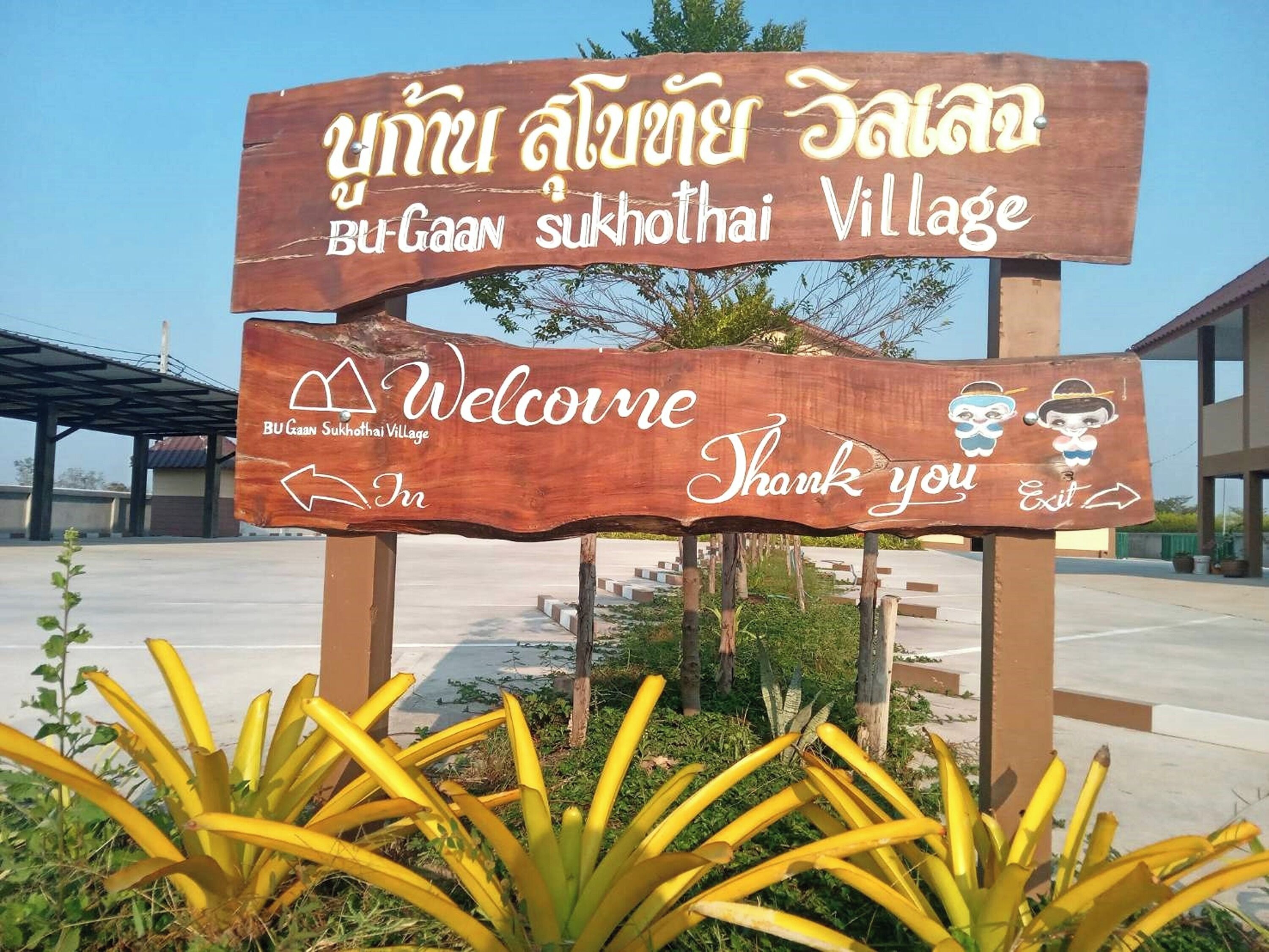 A wooden welcome sign for Bu Gaan Sukhotai Village with directional arrows and a 'Thank you' message.