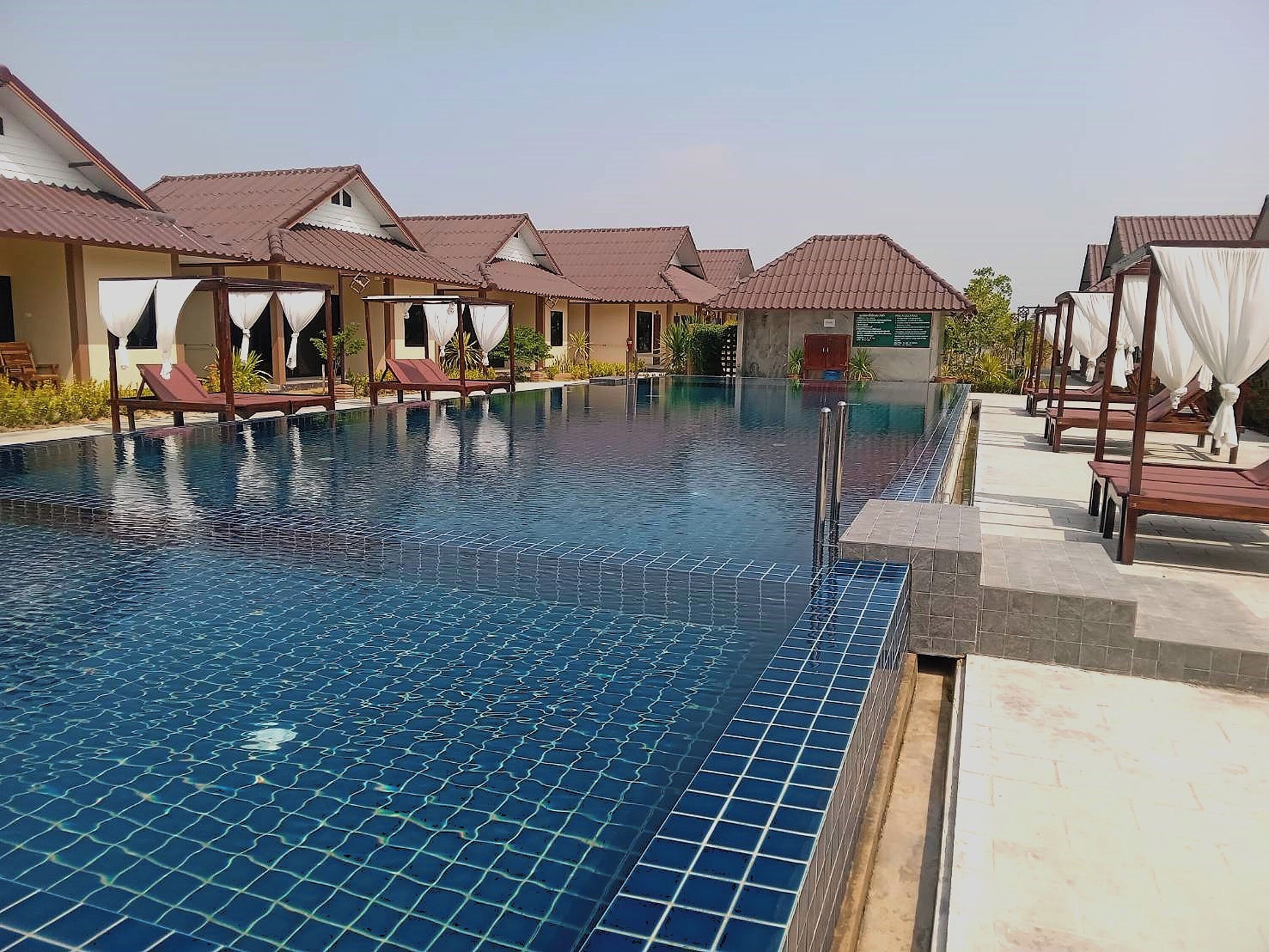 A swimming pool with lounge chairs and covered seating areas, surrounded by buildings with tiled roofs.