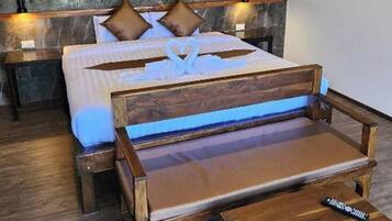 Una cama con una decoración con forma de corazón en las almohadas, un banco de madera y un control remoto sobre el banco.
