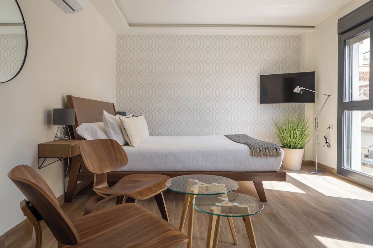 A modern hotel room with a bed, a desk, a chair, a small table, a TV, and a potted plant.