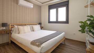 Un dormitorio con cama de madera, ropa de cama blanca, aire acondicionado de pared, ventana con persianas, mesita de noche con lámpara y planta en maceta.