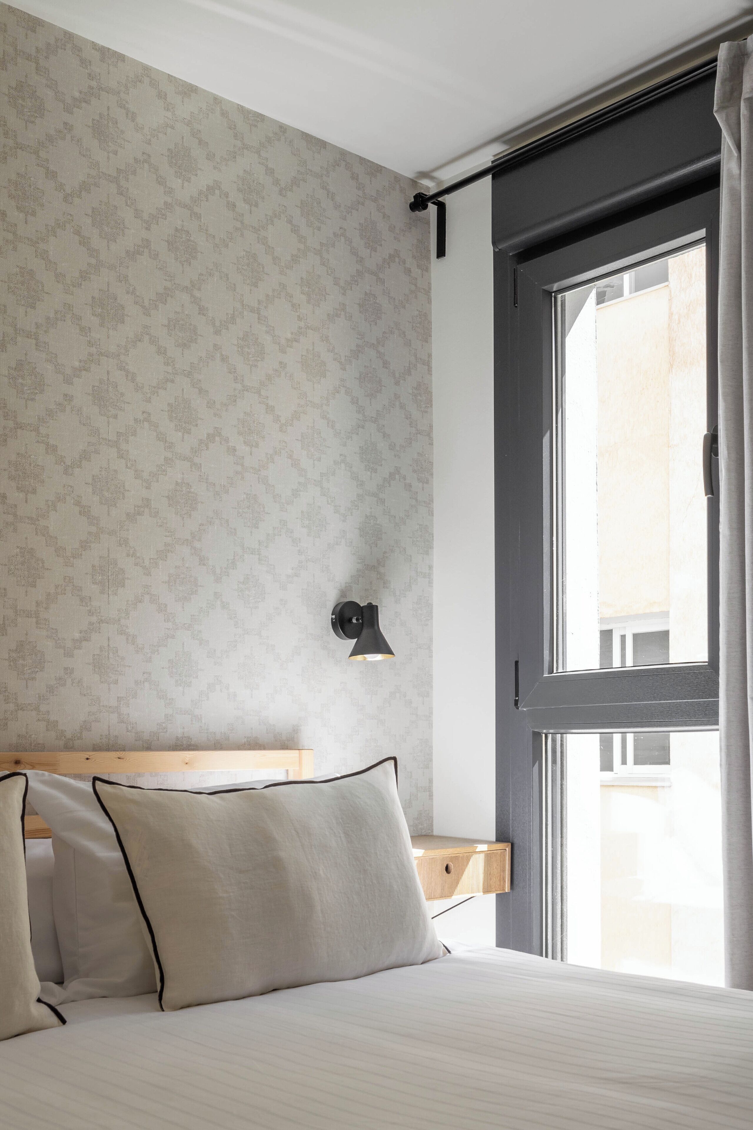 Una habitación de hotel con una cama, una mesita de noche, una lámpara de pared y un ventanal con cortinas.