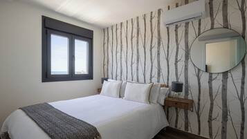A bedroom with a large bed, a window, a wall-mounted air conditioner, a round mirror, and a nightstand with a lamp.