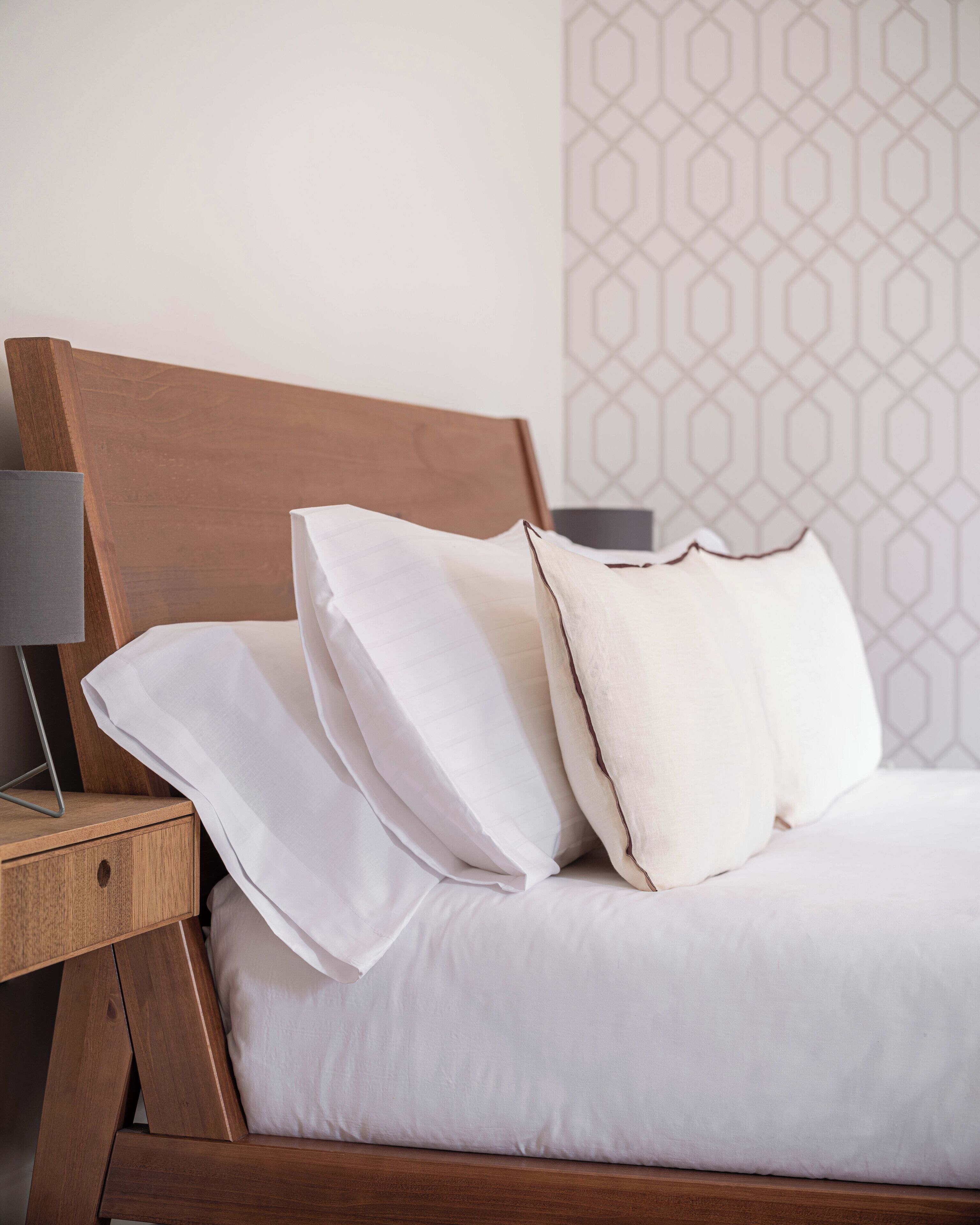 Een net opgemaakt bed met witte lakens en kussens, een houten hoofdbord en een nachtkastje met een lamp.