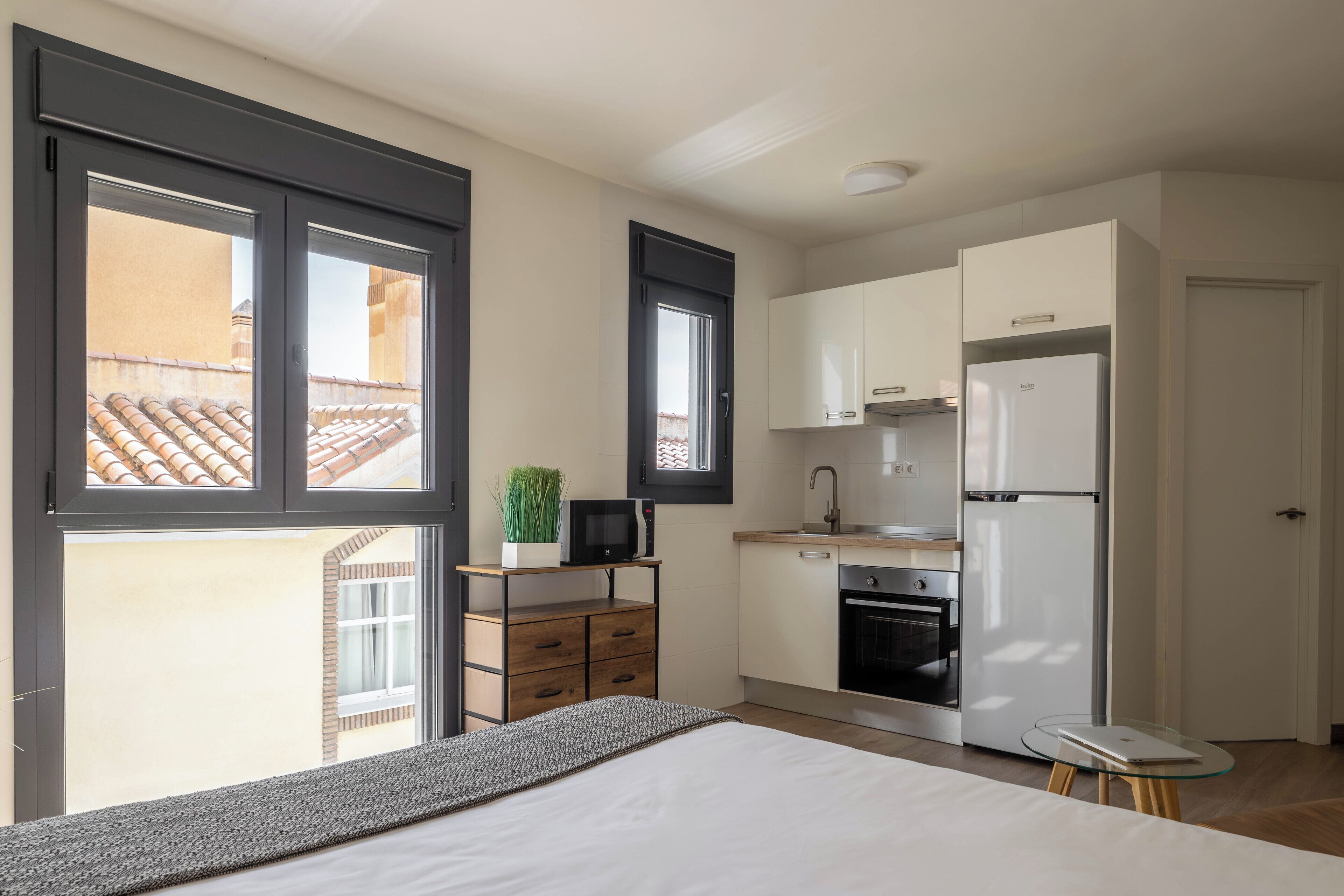 A compact kitchen with a refrigerator, microwave, and oven. There is a small table and a plant on a wooden shelf.