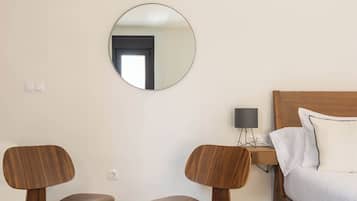 A modern hotel room with a wooden bed, a round mirror, a glass table, two wooden chairs, and a small desk with a lamp.