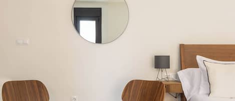 Una habitación de hotel moderna con una cama de madera, un espejo redondo, una mesita de cristal, dos sillas de madera y un escritorio pequeño con una lámpara.