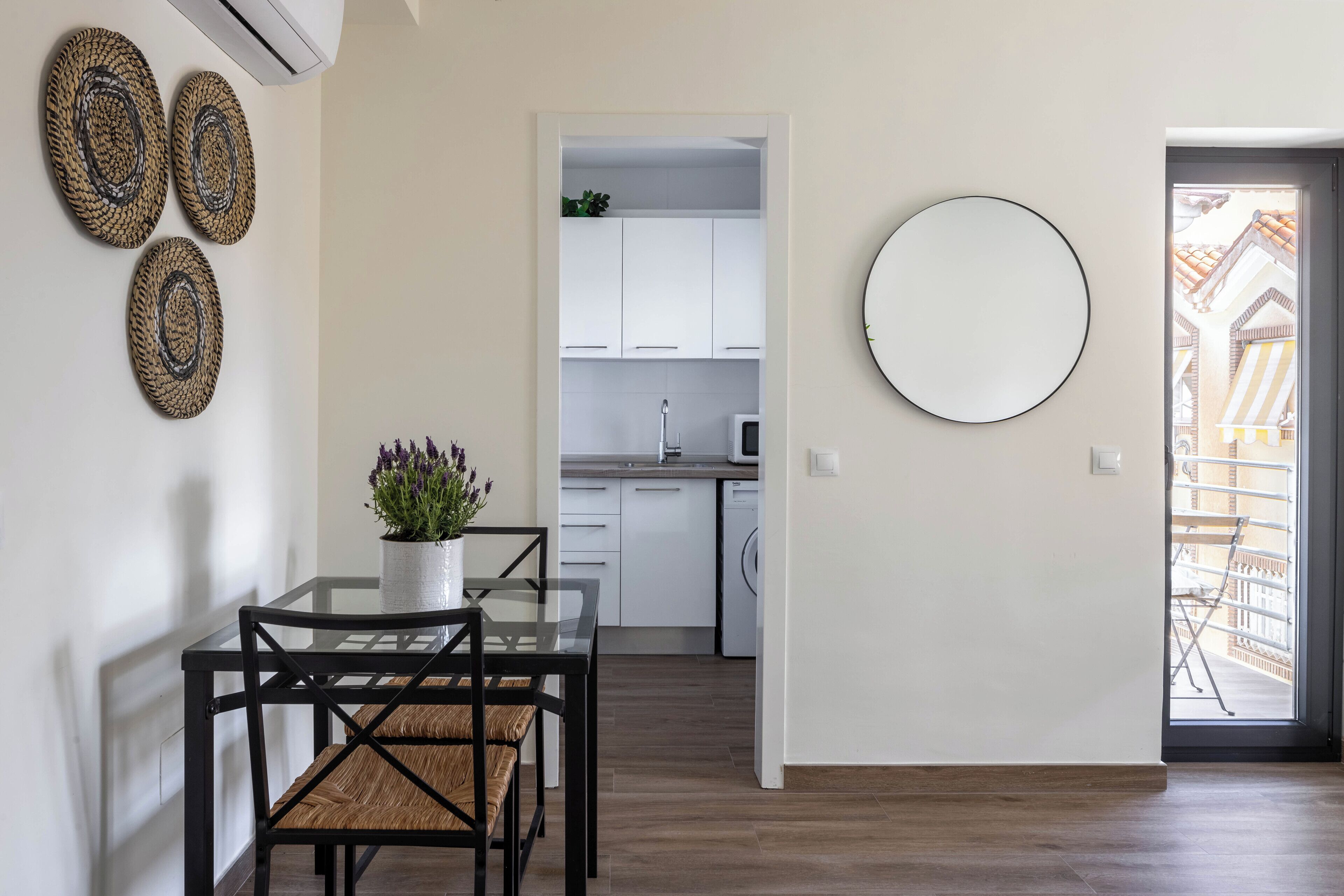 A modern kitchen with white cabinets, a dining area with a glass table and black chairs, and a round mirror on the wall.