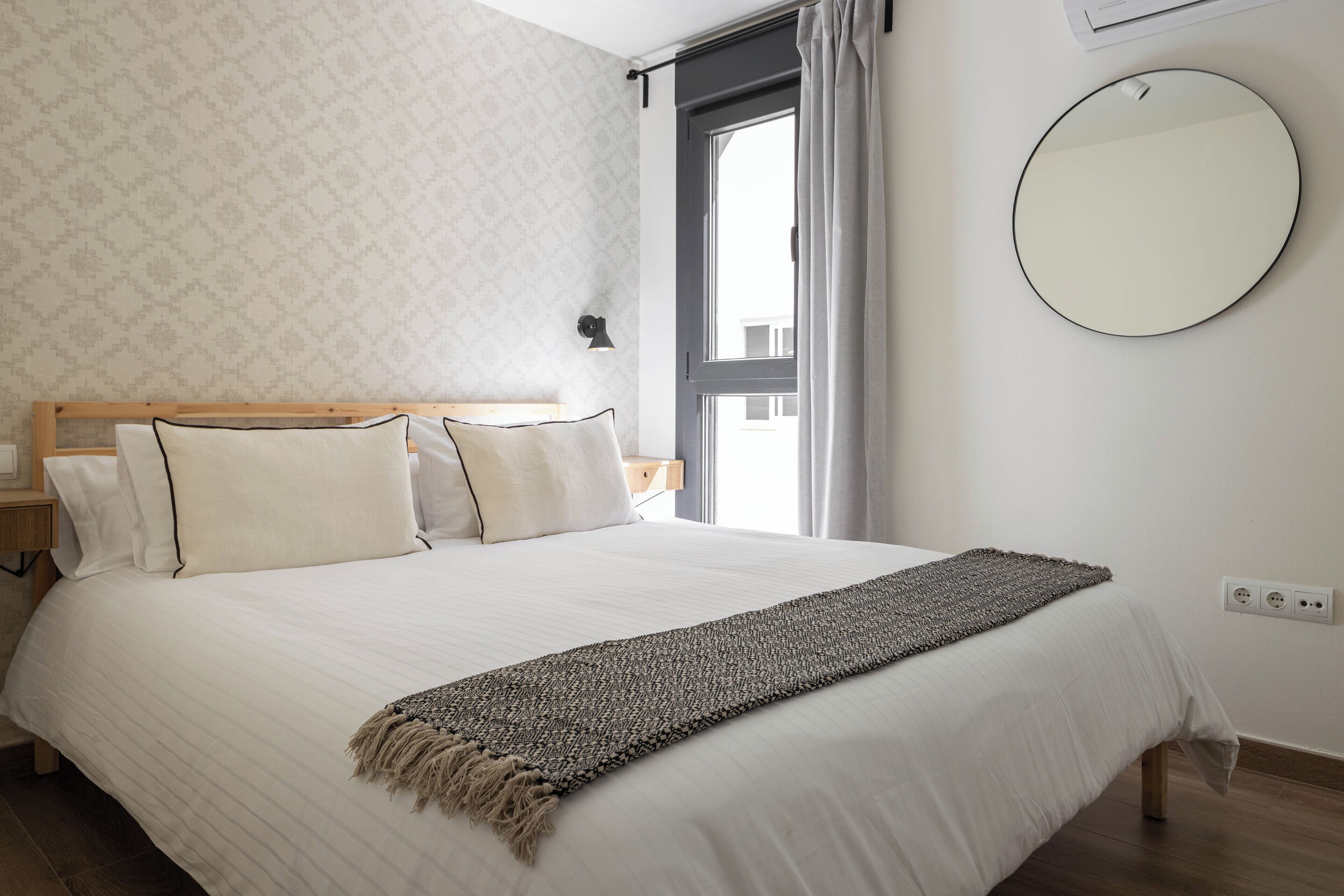 A neatly made bed with a white duvet and a textured throw blanket, a bedside table with a lamp, and a window with curtains.
