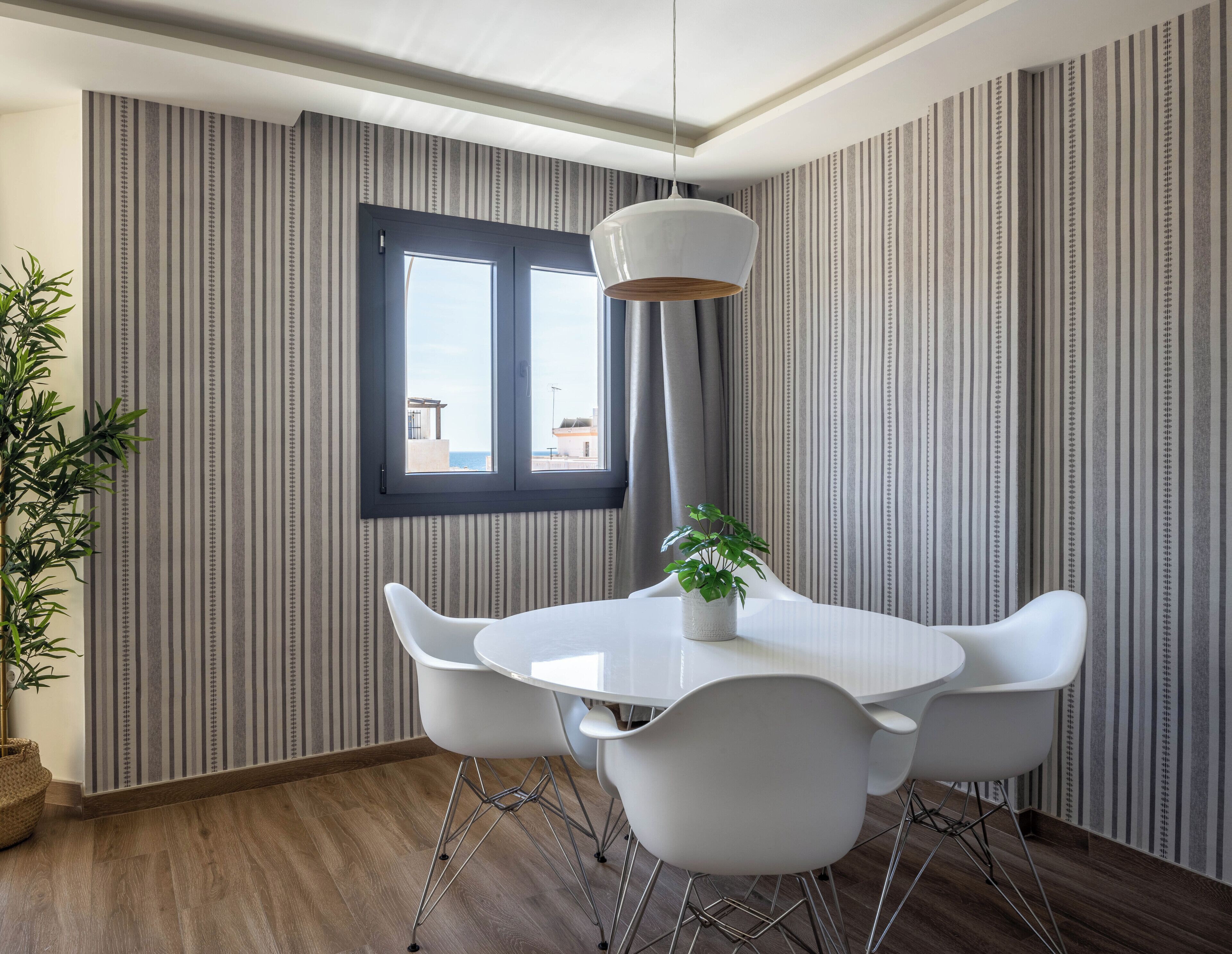 A modern dining area with a round table, white chairs, a hanging light fixture, and a window with a view.