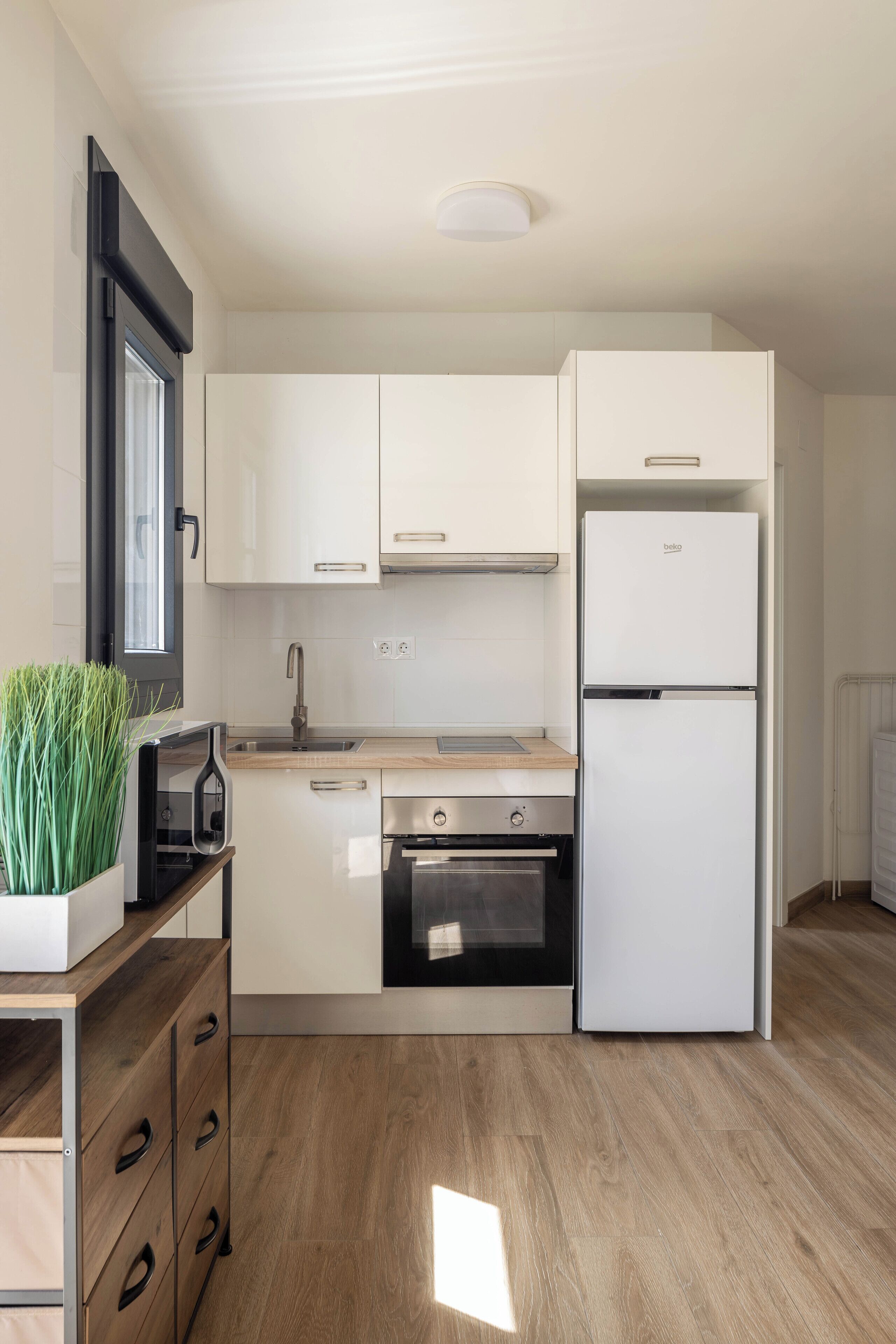 Una cocina moderna con armarios blancos, horno empotrado, refrigerador de dos puertas y ventana con marco negro.