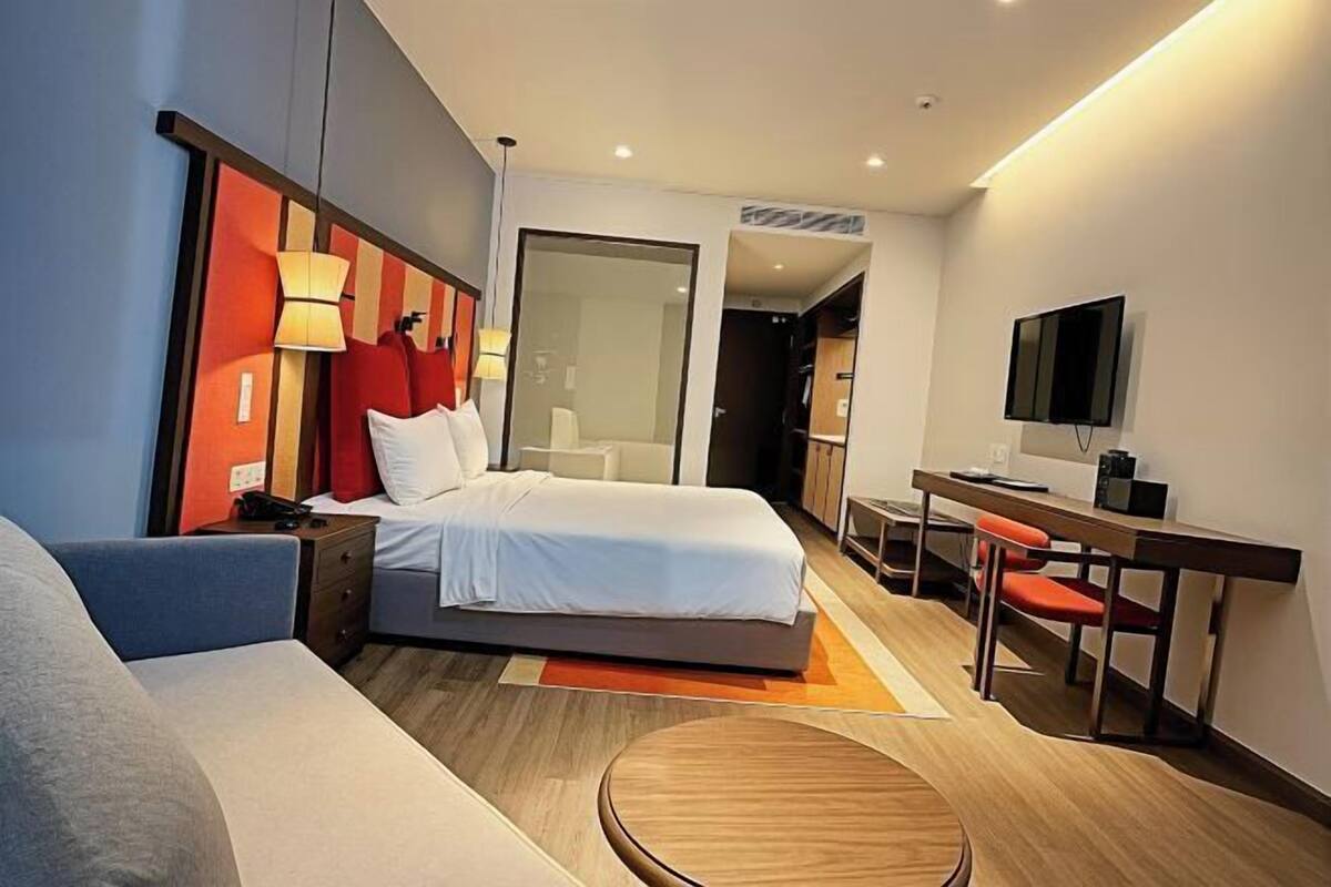 Une chambre d’hôtel moderne équipée d’un lit, d’une table de chevet, d’une lampe, d’un bureau avec une chaise, d’une télévision et d’une table ronde en bois.