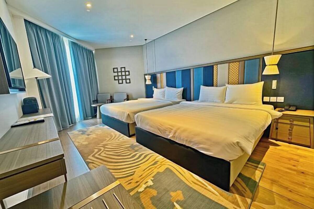 Une chambre d’hôtel avec deux lits, un bureau, une chaise, une lampe, une table de chevet et un tapis à motifs.