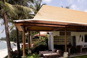 A covered outdoor seating area with a sofa and dining table, surrounded by palm trees and a view of the sea.