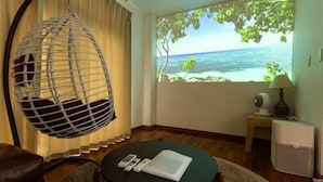A room with a hanging chair, a round table with a laptop, a projector screen displaying a tropical beach scene, and a small table with a lamp and a fan. - Mizunowa (Nago)