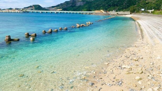 A beach with clear water, pebbles, and a pier in the background. - Mizunowa (Nago)